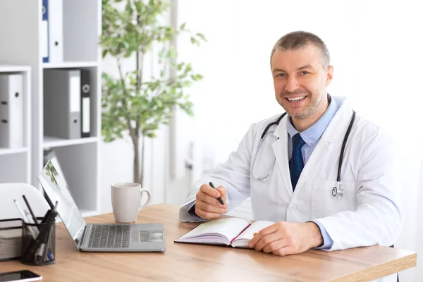 Portrét usmívajícího se doktora, jak se dívá na kameru sedícího u stolu — Stock fotografie