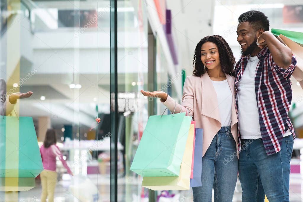 Discount Prices. Smiling Black Couple Shopping In Mall Together, Pointing At Showcase