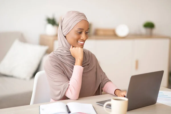 Mulher muçulmana negra feliz no hijab trabalhando remotamente no laptop de casa — Fotografia de Stock