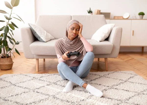Teljes hosszúságú portré feldúlt fekete nő hidzsáb elvesztése számítógépes játék, ül joystick a padlón otthon — Stock Fotó