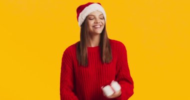 Noel Baba şapkalı çekici genç kadın Noel hediyesini sallıyor ve tatlı tatlı gülümsüyor, turuncu stüdyo geçmişi var.
