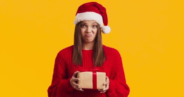 Joven mujer disgustada en Santa sombrero sosteniendo presente caja y frunciendo el ceño, fondo de estudio naranja — Vídeos de Stock