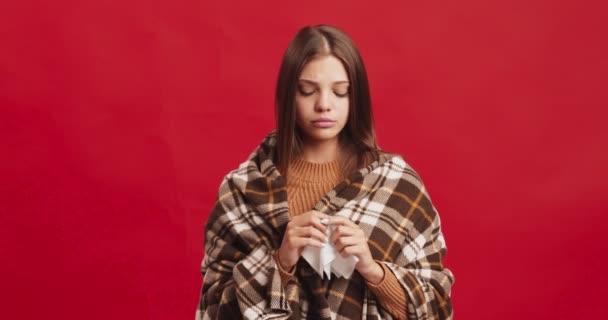 Young lady wrapped in plaid sneezing, blowing her nose into tissue, got sick before holidays, red studio background — Stock Video