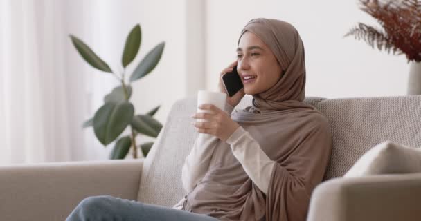 Ung muslimsk kvinde taler på mobiltelefon og nyder varm te, sidder på sofaen derhjemme, slow motion – Stock-video