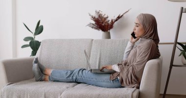 Evde modern işler. Laptop 'ta çalışan ve telefonla konuşan genç İslami bayanın yan görüntüsü.
