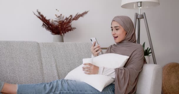 Joven mujer musulmana surfeando en las redes sociales en el teléfono móvil, disfrutando de las noticias, sentado con la taza de café en el sofá en casa — Vídeos de Stock