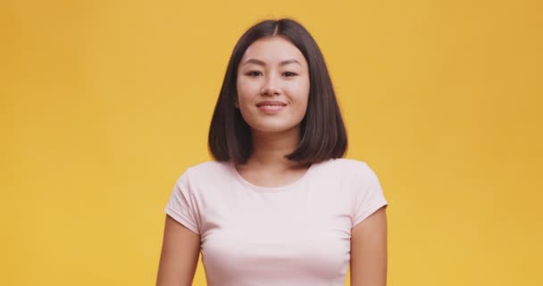 Sociável jovem asiático mulher sorrindo amigável para a câmera e acenando mão gesturing Olá, fundo amarelo — Vídeo de Stock