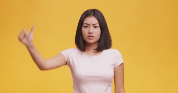 Концепция не нравится. Молодая недовольная азиатка жестикулирует большим пальцем вниз, оранжевый фон — стоковое видео