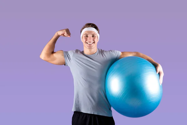 Feliz atleta milenario sosteniendo la pelota de fitness y mostrando sus bíceps en el fondo del estudio violeta — Foto de Stock