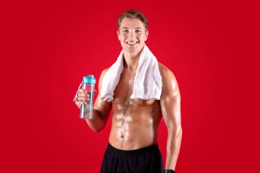 Egzersiz sırasında susuz kalma. Kırmızı stüdyo arka planında havlusu ve bir şişe suyu olan mutlu üstsüz sporcu.
