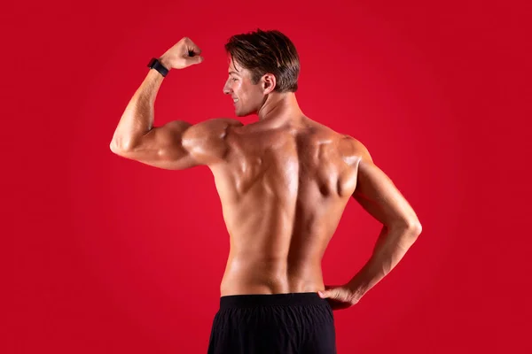 Rückenansicht des glücklichen jungen Bodybuilders, der seinen Bizeps über rotem Studiohintergrund zeigt — Stockfoto