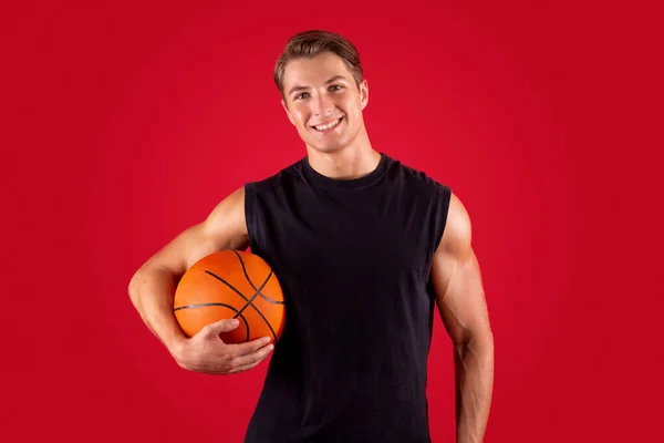 Portrait of happy young basketballer holding ball on red studio background — Stok Foto