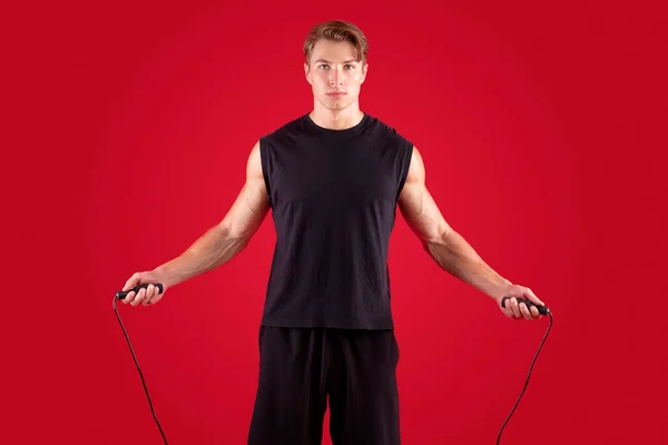 Aerobic workout concept. Serious athletic millennial guy jumping on skipping rope over red studio background — Stock Photo, Image
