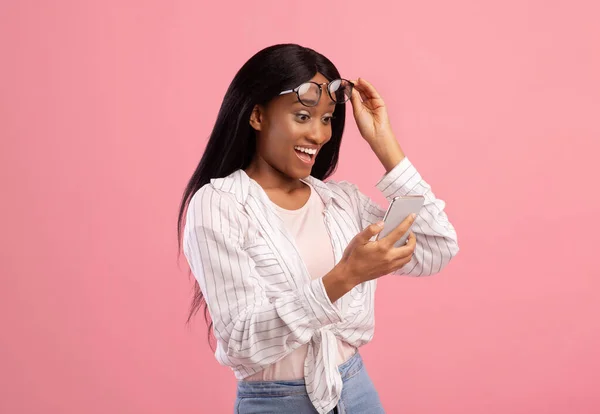 Millennial mulher negra em óculos olhando para o seu celular com surpresa sobre o fundo do estúdio rosa — Fotografia de Stock