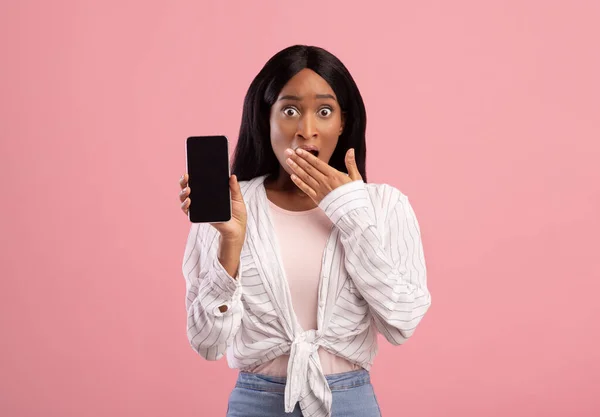 Zwarte dame toont smartphone met leeg scherm en het bedekken van haar mond in shock op roze achtergrond, mockup voor ontwerp — Stockfoto