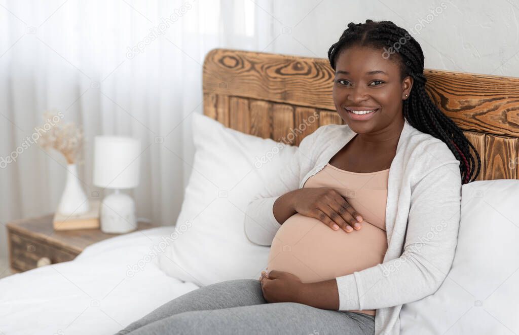 Smiling black expectant woman sitting on bed at home and touching belly