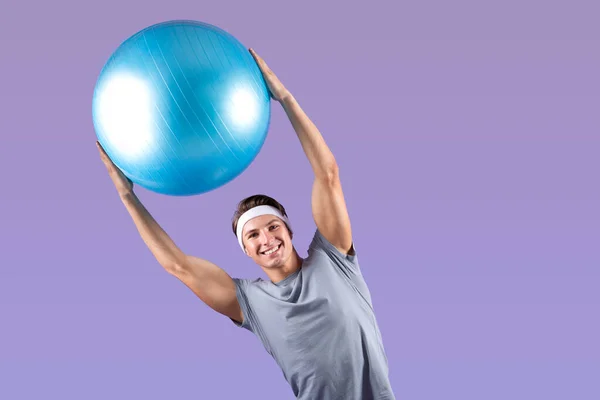 Позитивный молодой спортсмен поднимает над головой фитнес-мяч, откидываясь на фоне лиловой студии, копирует пространство — стоковое фото