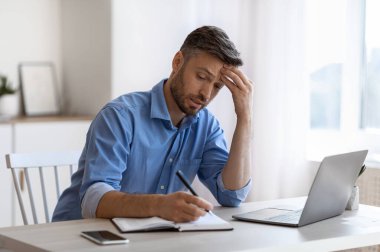 Ofiste dizüstü bilgisayarla çalışırken not almakla meşgul bir iş adamı.