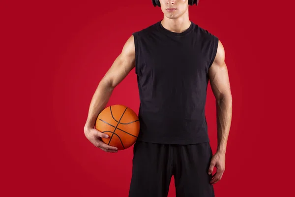 Vista cortada de jogador de basquete jovem segurando bola, ouvindo música em fones de ouvido sobre fundo vermelho, espaço de cópia — Fotografia de Stock