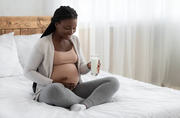 Кальцій під час вагітності. Чорна вагітна жінка сидить на ліжку тримає молоко скло — стокове фото