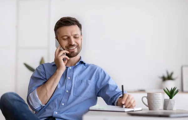 Hombre de negocios guapo hablando por celular y tomando notas mientras trabaja en la oficina — Foto de Stock