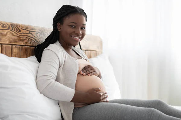 Glimlachende zwarte zwangere vrouw rusten op bed en het houden van handen op de buik — Stockfoto