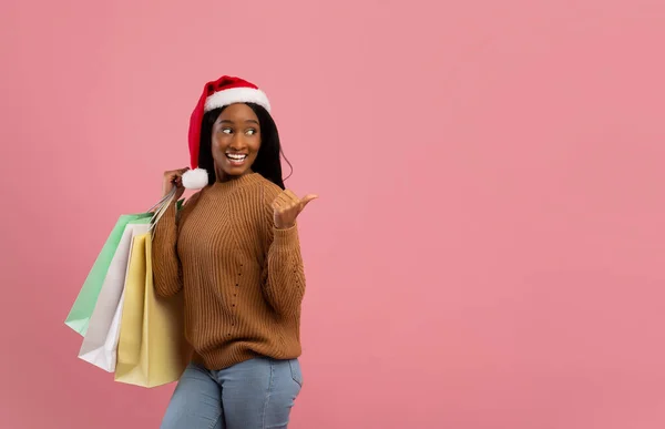 Enorme vendita di Natale. Attraente donna nera in cappello Babbo Natale in possesso di sacchetti regalo e indicando lo spazio vuoto, sfondo rosa — Foto Stock