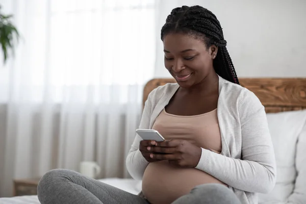 Програма для стеження за вагітністю. Щаслива чорна вагітна жінка використовує смартфон вдома — стокове фото