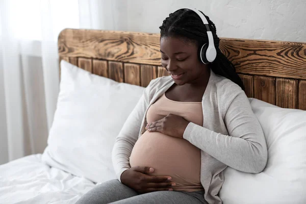 Музыка для беременных. Счастливая чернокожая беременная женщина в беспроводных наушниках, расслабляющихся на кровати — стоковое фото