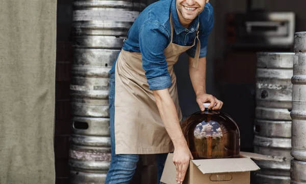 Sorrindo bonito jovem trabalhador homem no avental tira grande frasco para cerveja e bebidas de produção — Fotografia de Stock