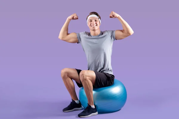 Positiv ung idrottsman sitter på fitness boll och visar sin styrka på lila studio bakgrund — Stockfoto