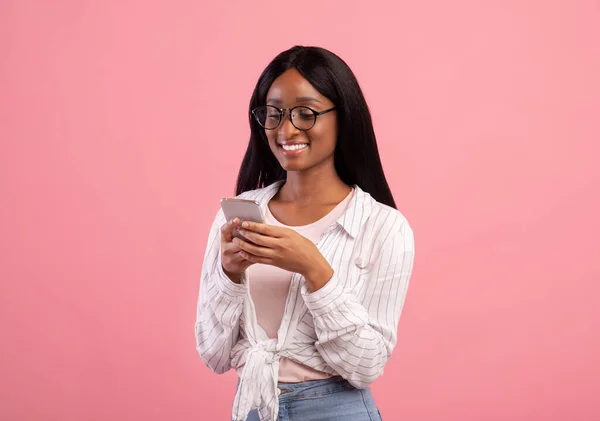 Vida en línea. Mujer negra inteligente en gafas usando teléfono celular para trabajo remoto o estudios sobre fondo de estudio rosa —  Fotos de Stock