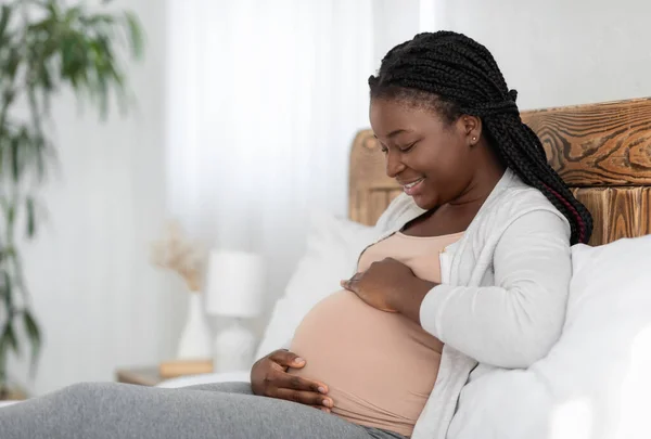Vrolijke Afro-Amerikaanse zwangere vrouw ontspannen op bed thuis, aanraken buik — Stockfoto