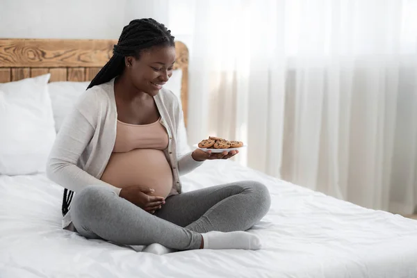 Mujer embarazada feliz con plato de galletas sentado en la cama en casa — Foto de Stock