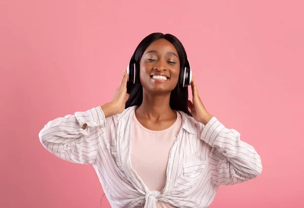 Mulher negra relaxada em fones de ouvido desfrutando de boa música e sorrindo com os olhos fechados no fundo do estúdio rosa — Fotografia de Stock