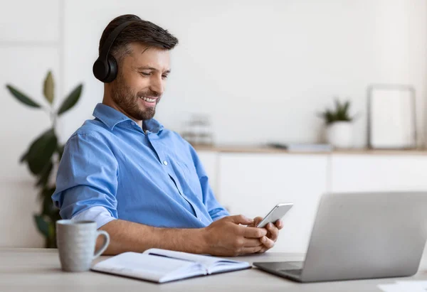 Hombre de negocios guapo escuchando música en el teléfono inteligente con auriculares mientras trabaja en la oficina —  Fotos de Stock
