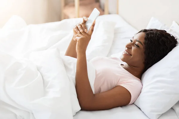 Mujer negra alegre acostada en la cama, usando el teléfono móvil — Foto de Stock