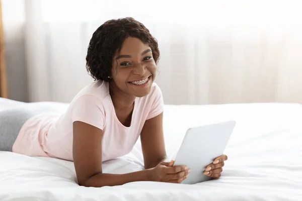Yatakta dijital tablet kullanan gülümseyen siyah kadın. — Stok fotoğraf