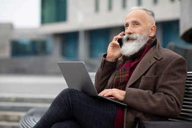 Dizüstü bilgisayarı olan yaşlı işadamı bankta oturuyor, telefonla konuşuyor.