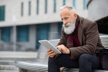 Beyaz saçlı şık adam bankta oturuyor, dijital tablet kullanıyor.