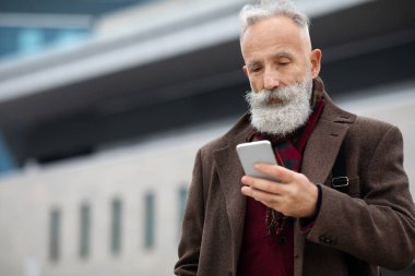 Gri saçlı işadamı sokakta akıllı telefon kullanıyor.