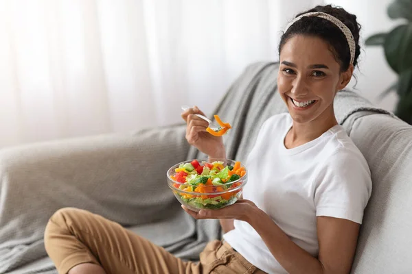 Ung kvinna sitter på soffan hemma, äter hälsosam mat — Stockfoto