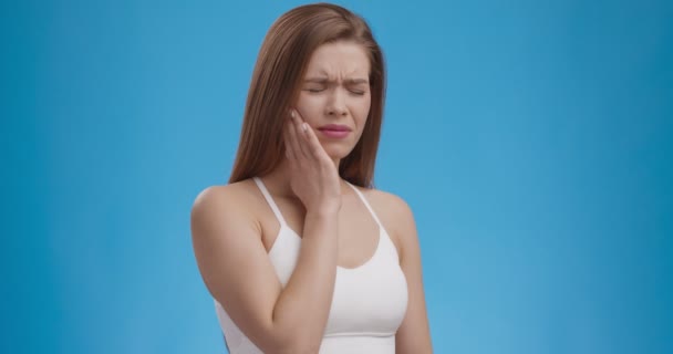 Junge Frau leidet unter akuten Zahnschmerzen, berührt ihren schmerzenden Kiefer und runzelt die Stirn, blauer Studiohintergrund — Stockvideo