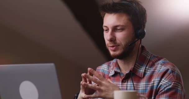 Pewny siebie facet w słuchawkach rozmawiający przez internet na wideokonferencji, wyjaśniający projekt gestami w odległym miejscu pracy — Wideo stockowe