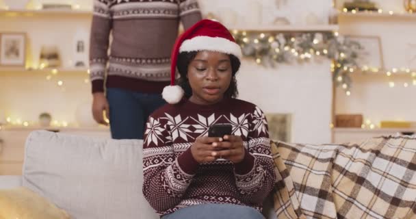 Mujer negra trabajando en red en el teléfono celular, hombre sorprendiéndola, cerrando los ojos de la mujer. Celebración de Navidad juntos concepto — Vídeo de stock