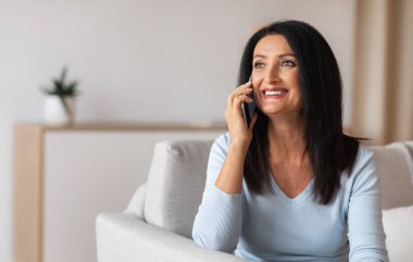 Gülümseyen olgun kadın koltukta oturmuş cep telefonuyla konuşuyor.
