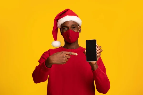 Afrikanischer Kerl mit Maske und Weihnachtsmütze zeigt auf schwarzen Smartphone-Bildschirm — Stockfoto