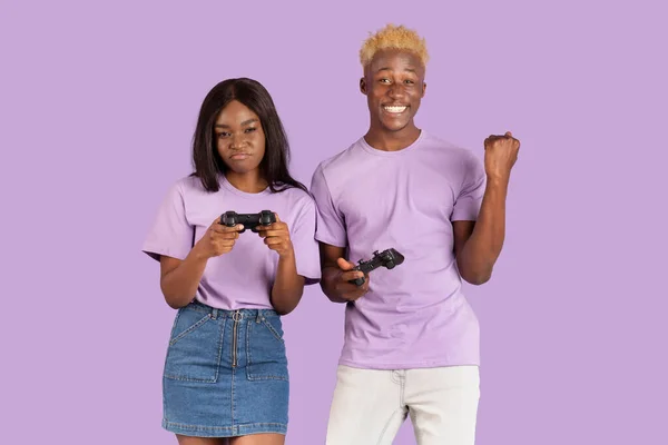 Glücklicher afroamerikanischer Typ mit Joystick gewinnt Videospiel über seine aufgebrachte Freundin auf violettem Studiohintergrund — Stockfoto