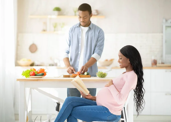 Afro-americano expectante mulher ler livro enquanto seu marido cozinhar salada de legumes na cozinha — Fotografia de Stock