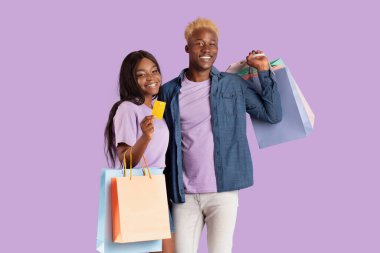 Menekşe stüdyosunun arka planında alışveriş torbaları ve kredi kartı olan genç Afrikalı Amerikalı erkek ve kız arkadaş